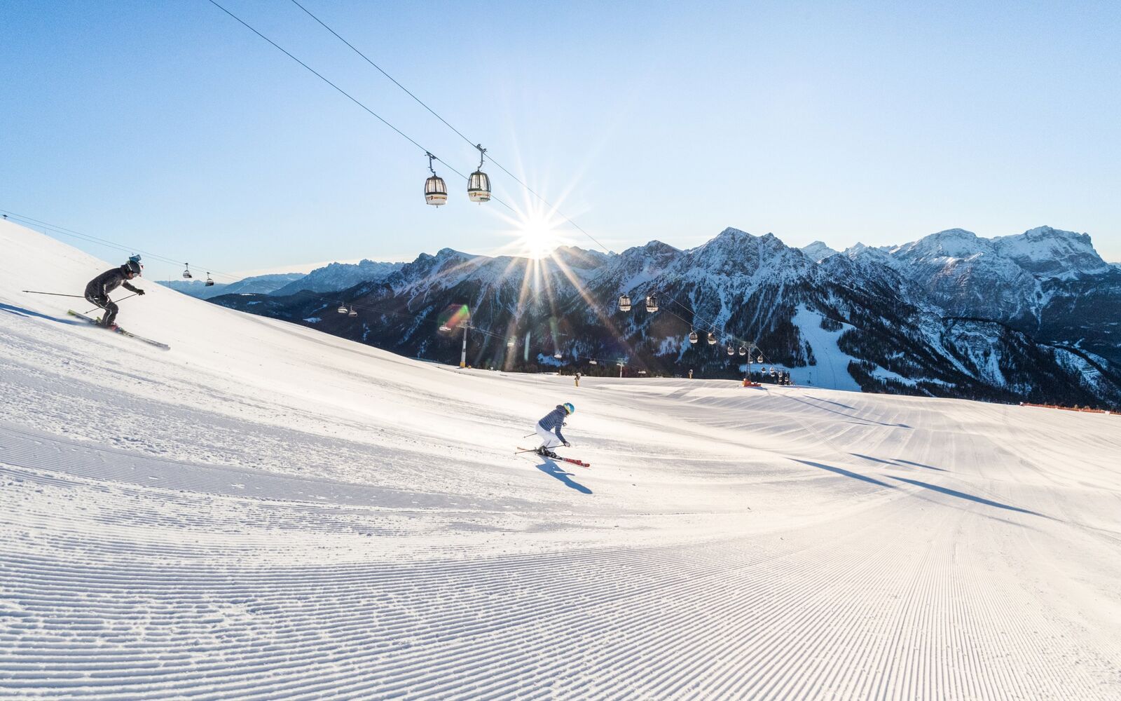 Zwei Skifahrer auf einer breiten, fast leeren Piste unter der Gondel am Kronplatz